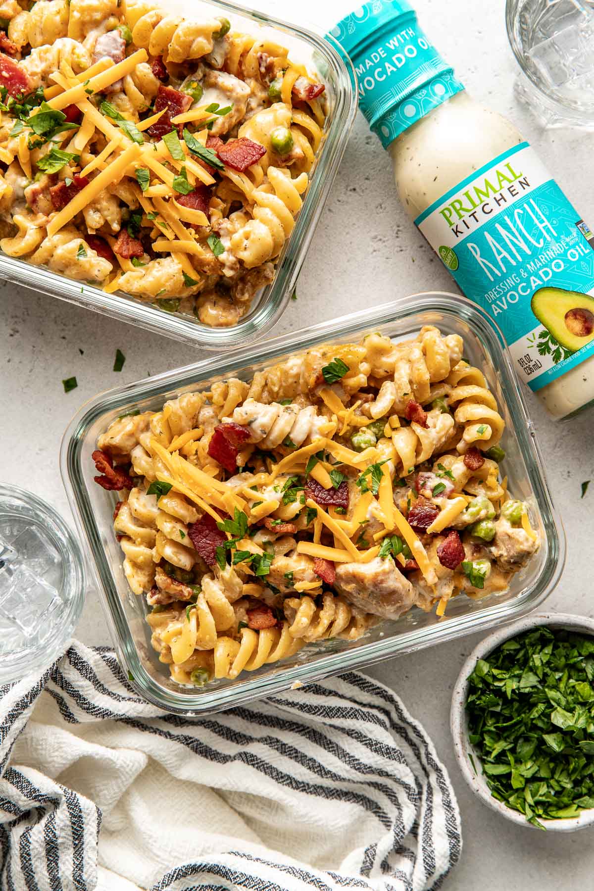 Overhead view of two glass meal prep containers filled with One-Pot Chicken Bacon Ranch Pasta and a bottle of primal kitchen ranch dressing on the side. 