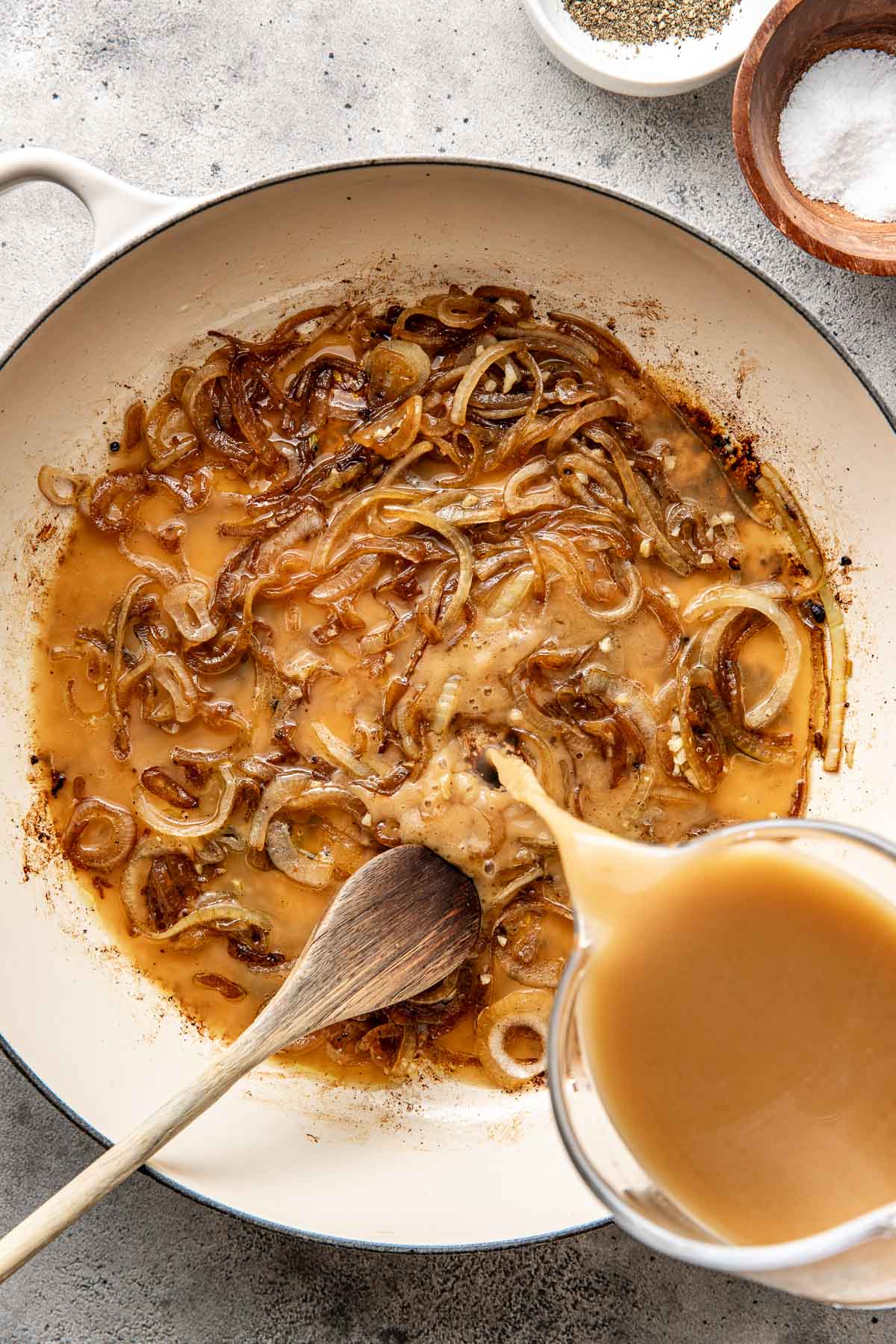 Overhead view of a skillet with a hand over it pouring a broth into an onion mixture. 
