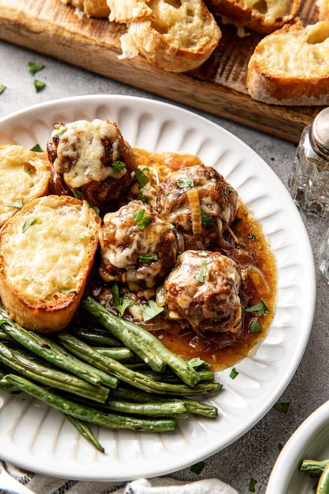 Close up view of a plate with French onion meatballs on it with green beans and a piece of bread on the side. 