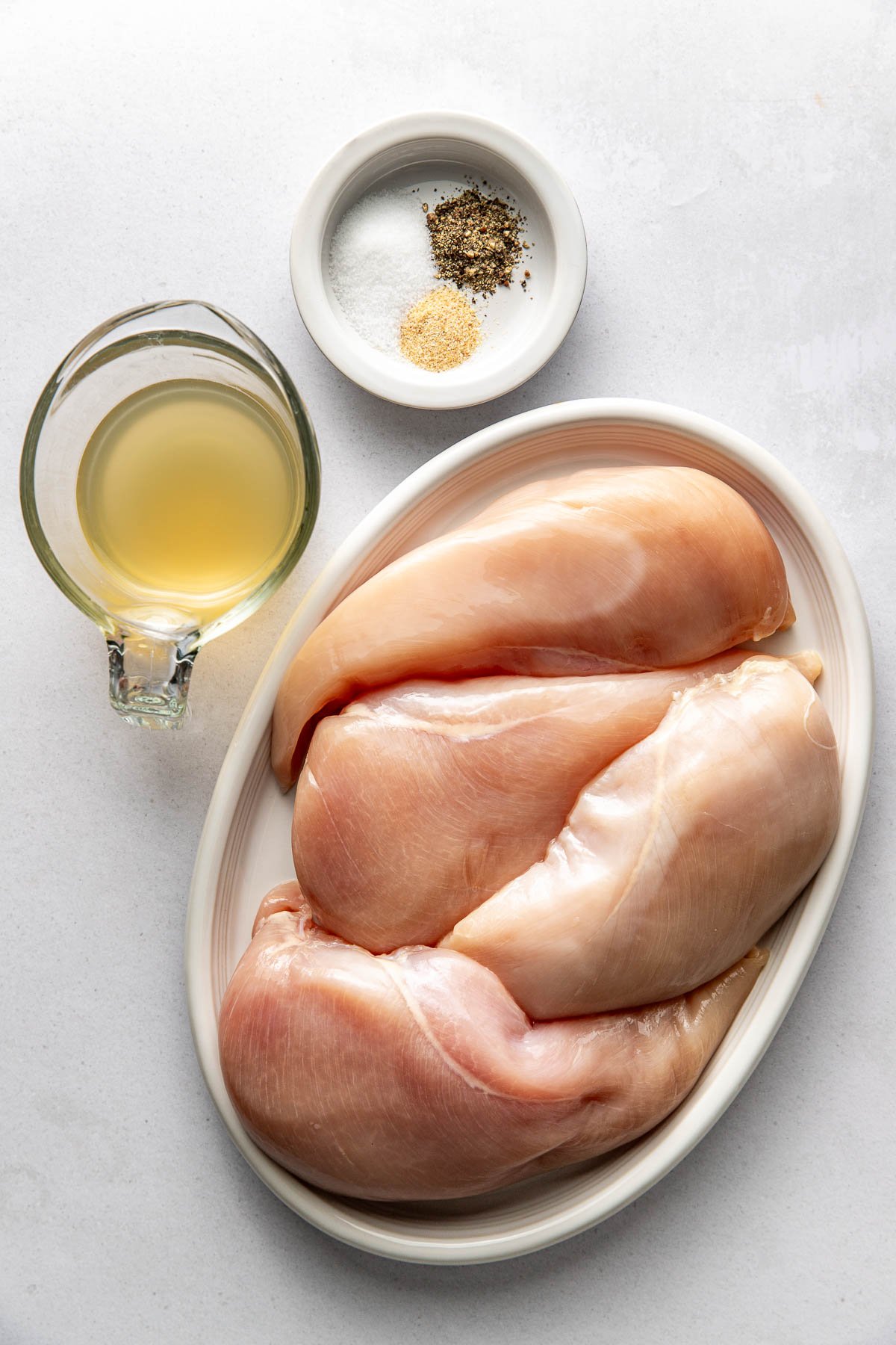 Overhead view of large plate filled with raw chicken breast next to a glass cup of broth and small dish of seasonings. 