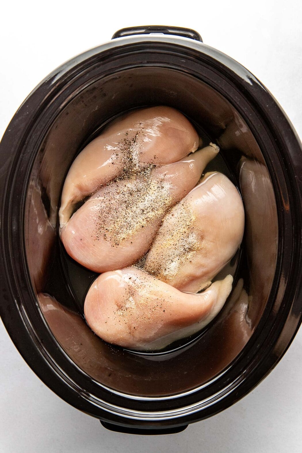 Overhead view of a slow cooker filled with four raw chicken breasts seasoned on top.
