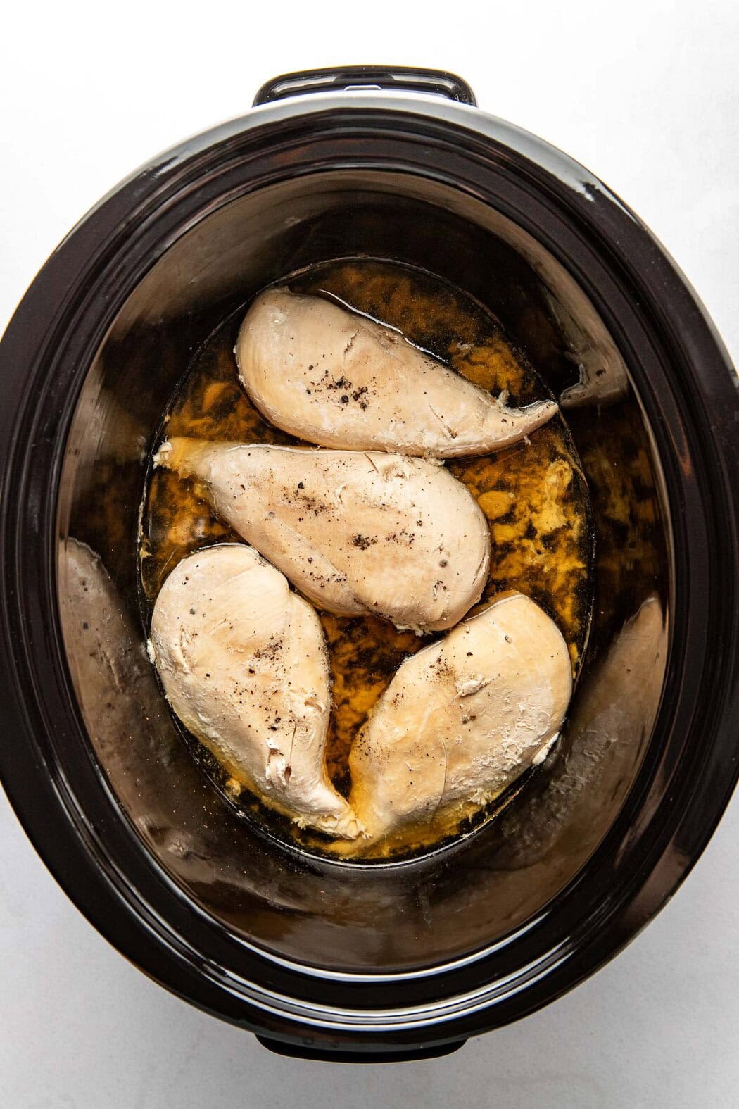 Overhead view of a slow cooker with freshly cooked chicken breasts in broth. 