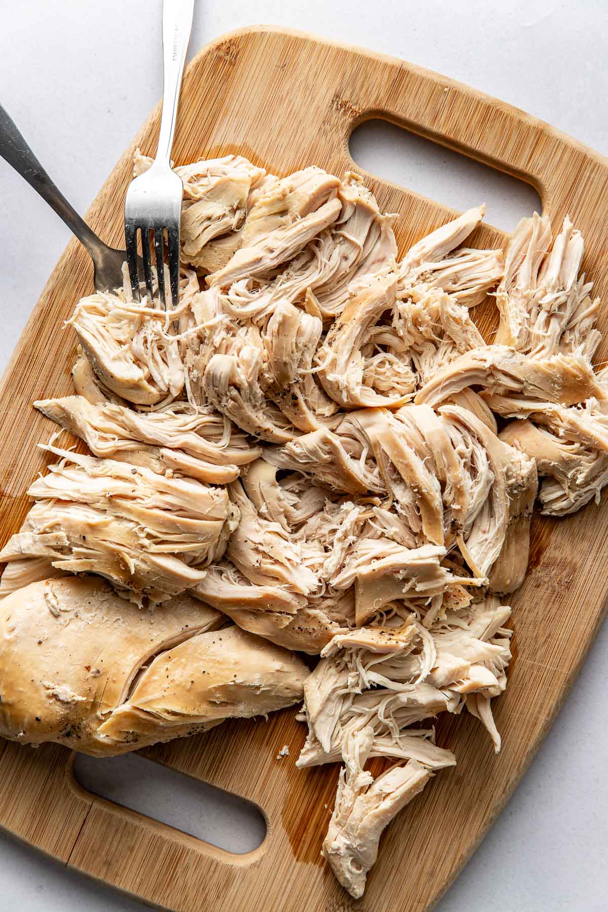 Overhead view of a wooden cutting board filled with shredded chicken and two forks. 