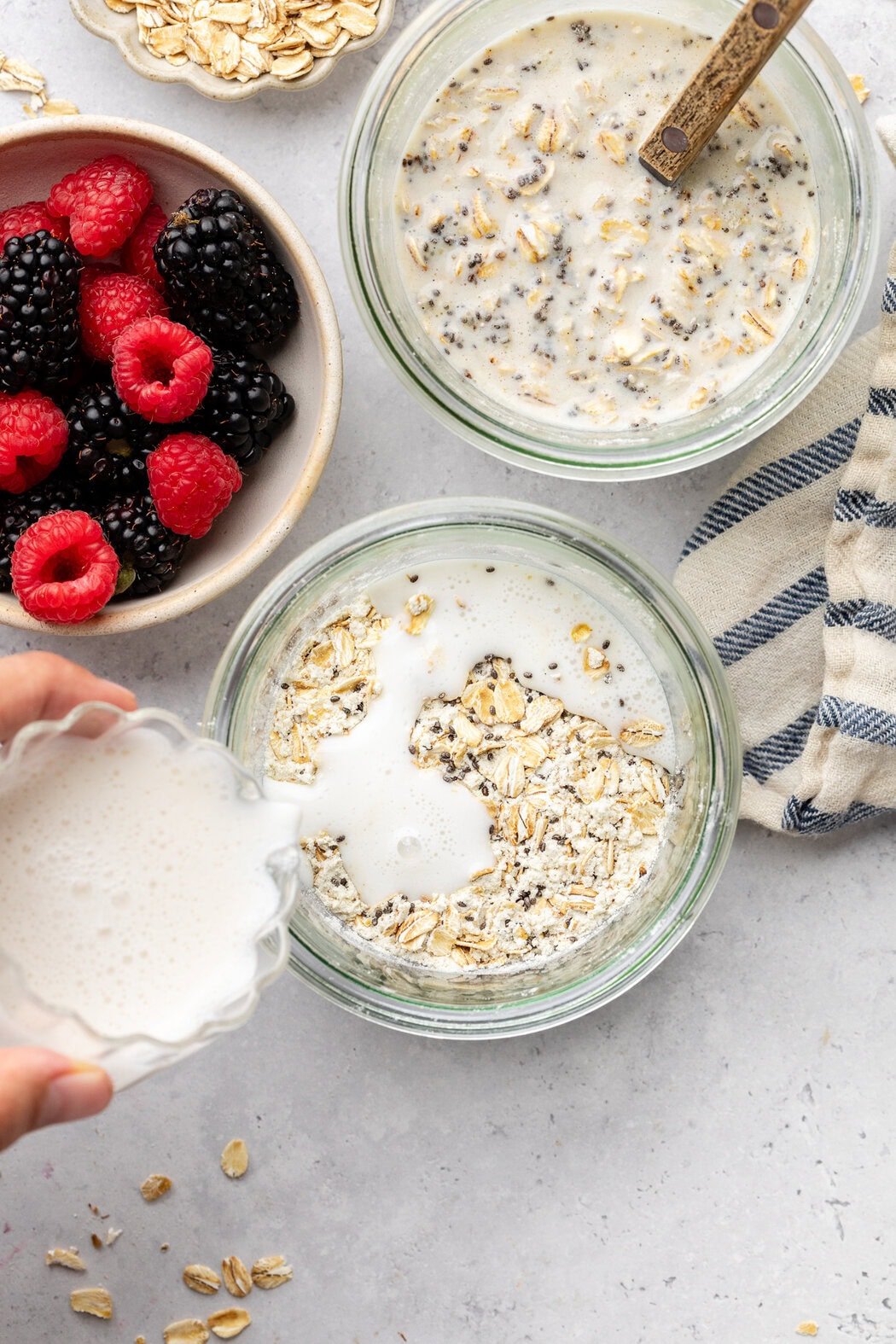 Overhead view of two glass jars filled with Protein Overnight Oats ingredients with a hand pouring milk on the top.