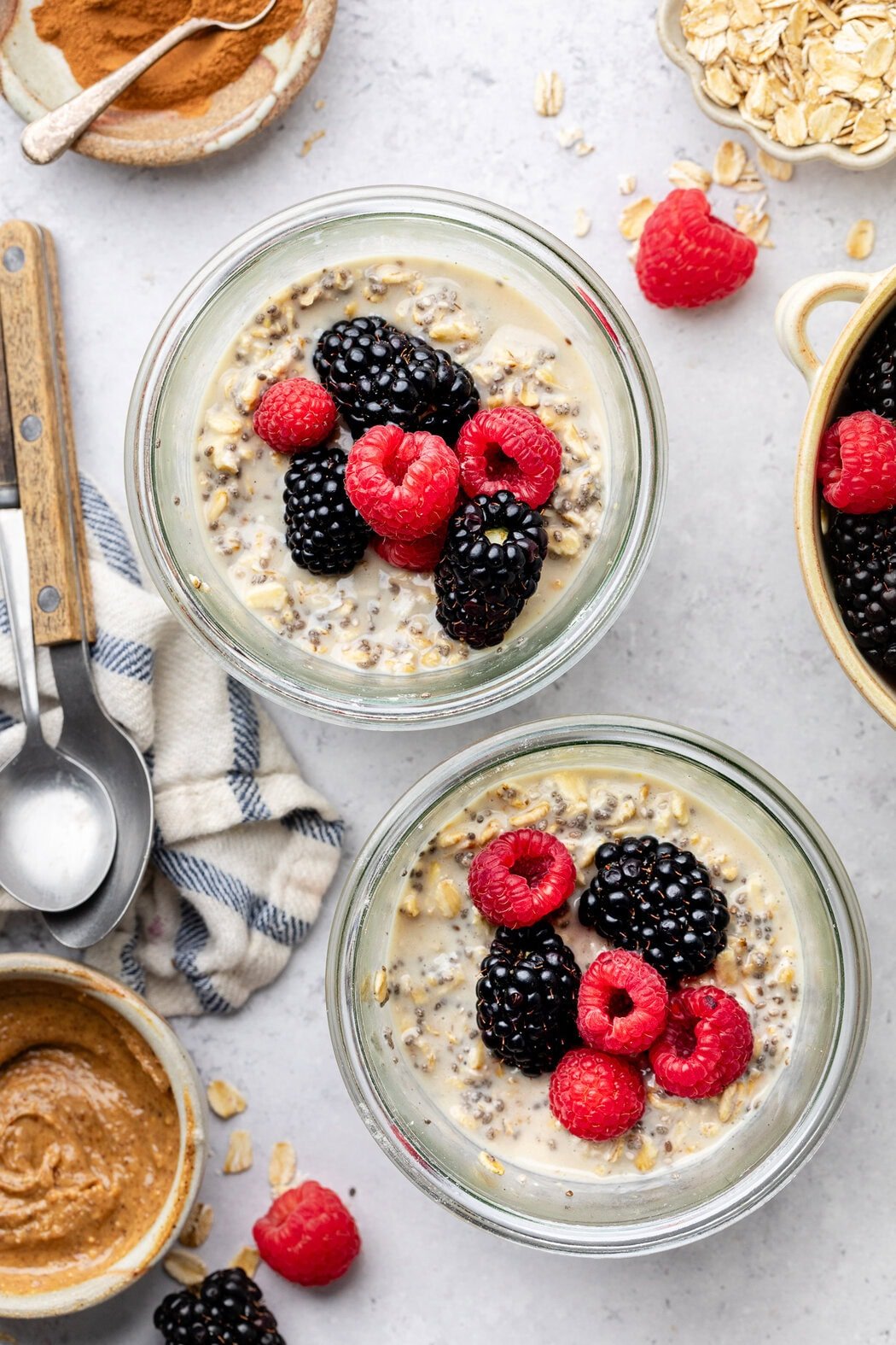 Overhead view of two jars of Protein Overnight Oats topped with colorful fresh berries and a bowl of nut butter on the side.
