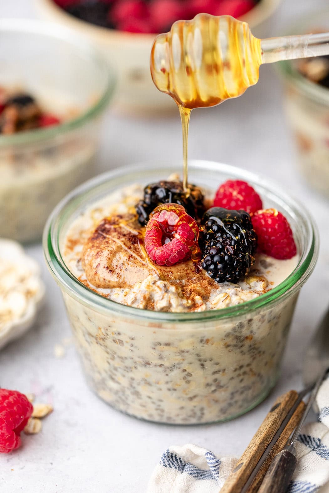 Close up view of honey being drizzled over a glass jar of Protein Overnight Oats topped with fresh colorful berries. 