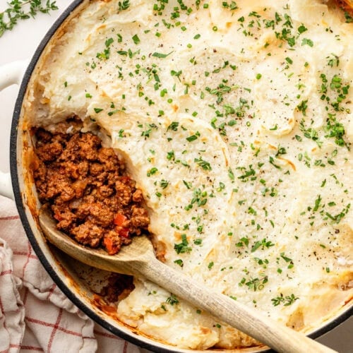 Overhead view of a round white casserole dish filled with mashed potato-topped sloppy joe mixture with a wooden spoon removing a portion.