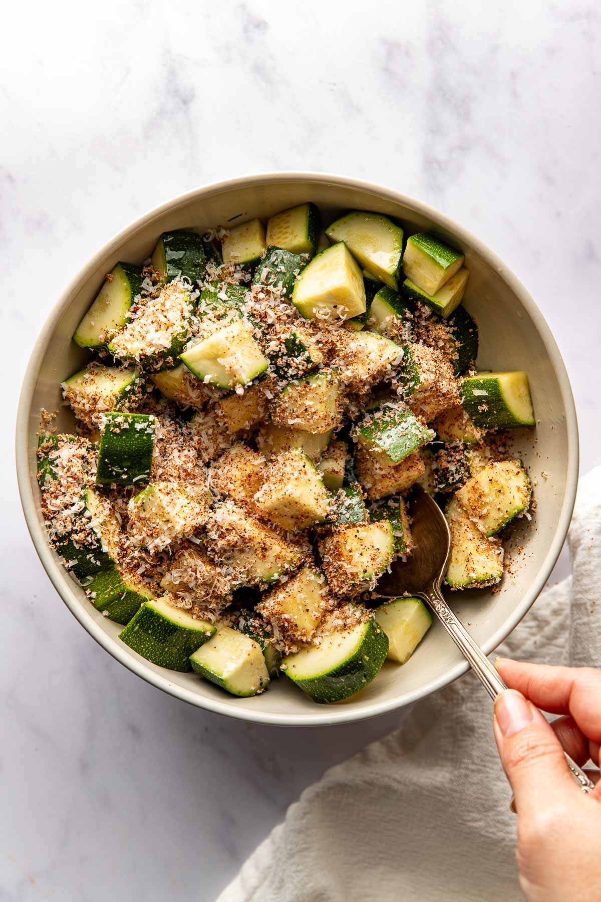 Overhead view of a bowl filled with zucchini pieces topped with seasonings and being stirred with a spoon. 