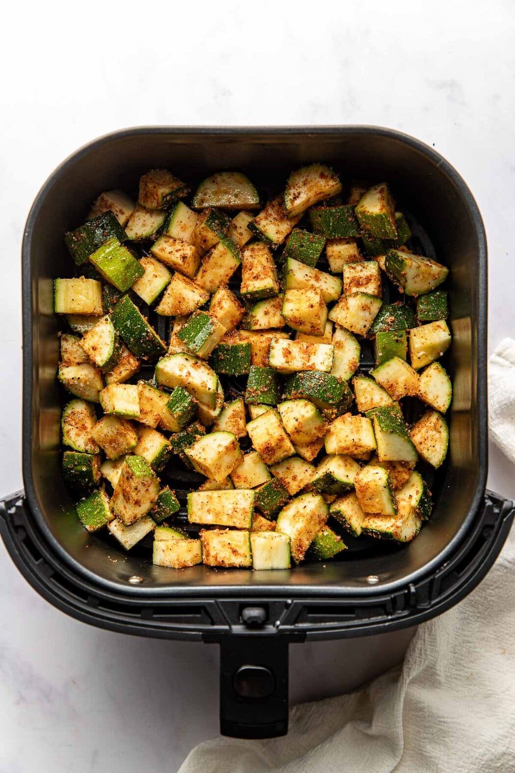 Overhead view of an air fryer basket filled with zucchini pieces coated in seasonings ready for roasting. 