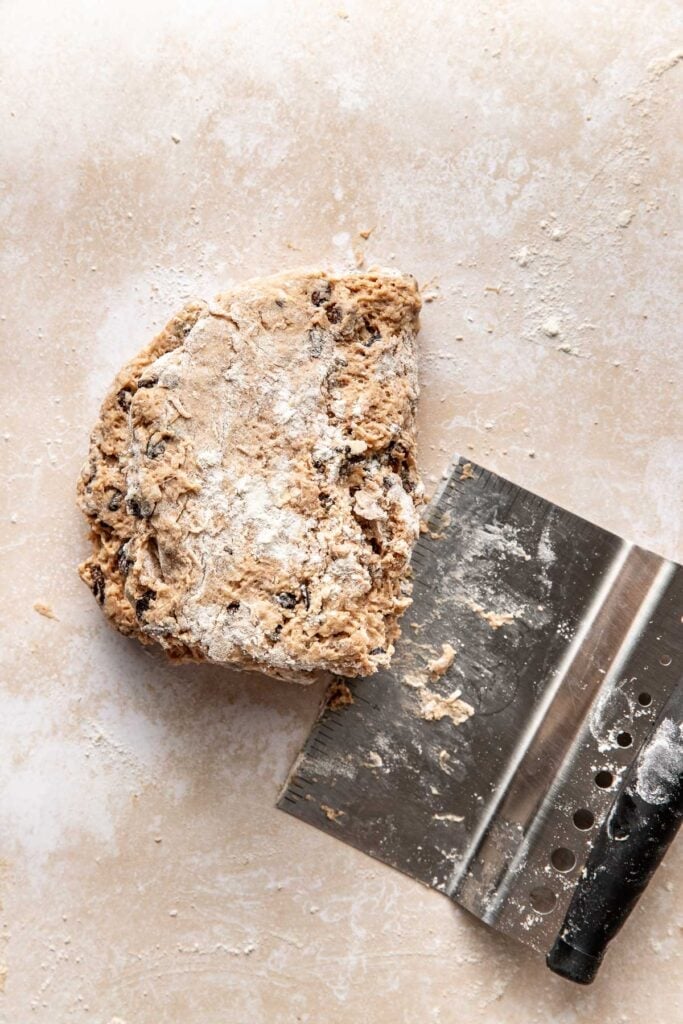 A piece of floury biscuit dough on the counter next to a metal bench scraper.