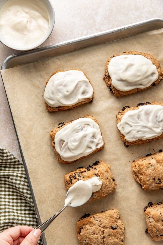 Overhead view of a sheet pan lined with parchment paper showing freshly baked Cinnamon Raisin Biscuits being topped with glaze. 