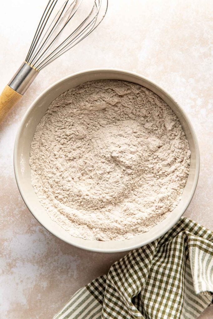 A large white mixing bowl filled with flour next to a wire whisk with a wooden handle