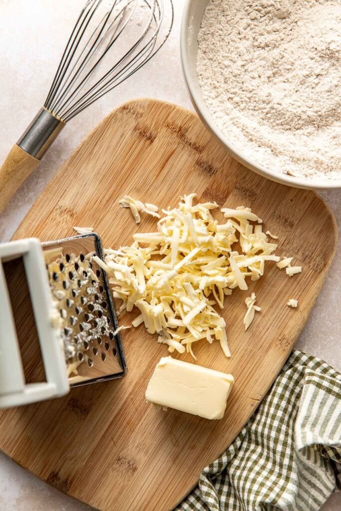 A white-handled box grater on a wooden cutting board with shreds of bold butter that have been grated to make biscuits.