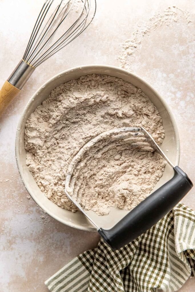 A pastry cutter lying along the side of a bowl filled with a coarse mixture of butter and flour.
