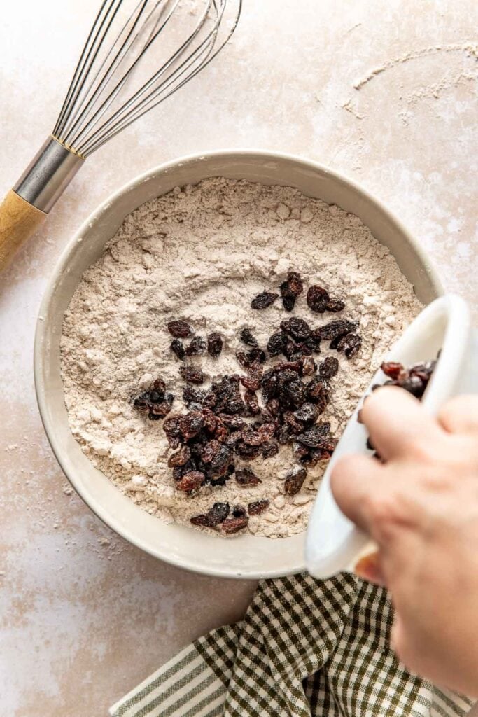 A hand pours raisins from a small white bowl into a bowl of dry ingredients for making biscuits.