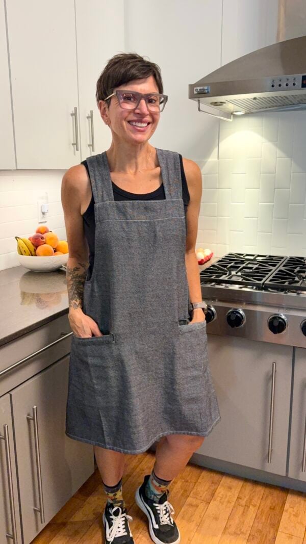 A woman with short dark hair and glasses wearing a grey smock-style apron in a kitchen.