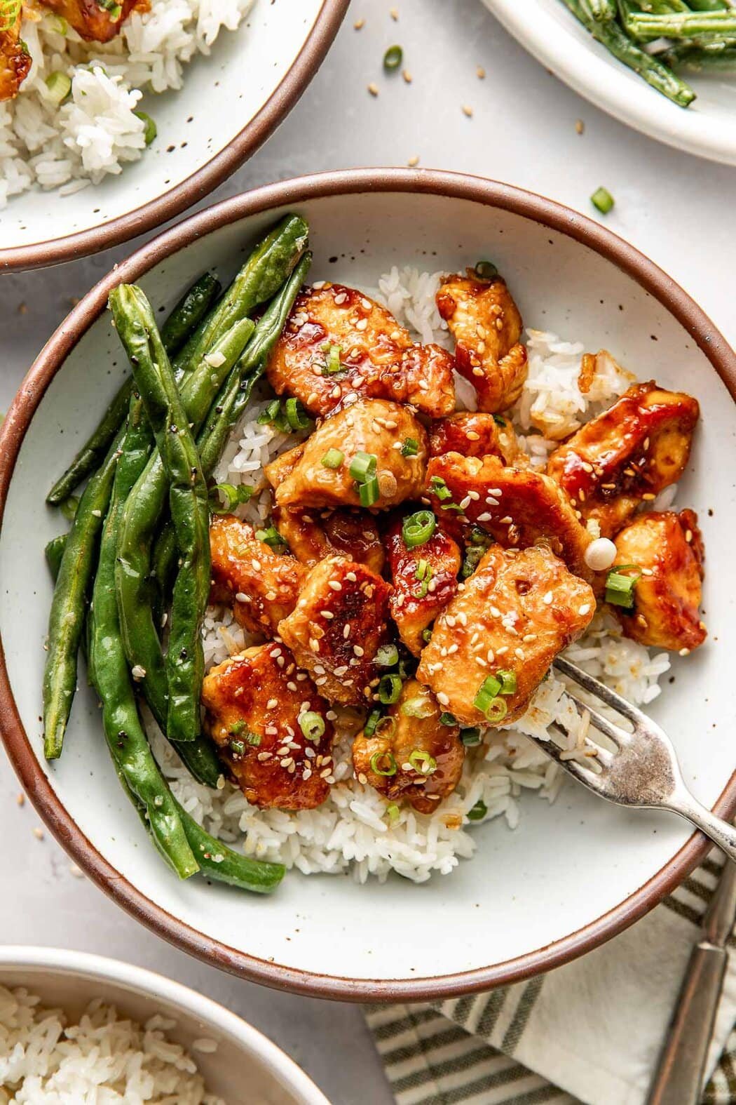 Overhead view of a plate of Sesame Chicken on top of white rice sprinkled with green onions next to roasted green beans. 