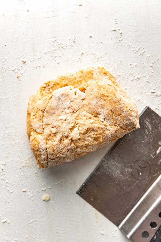 A stainless steel bench scraper lies next to a piece of sweet potato biscuit dough that has been folded in half on a floured surface.