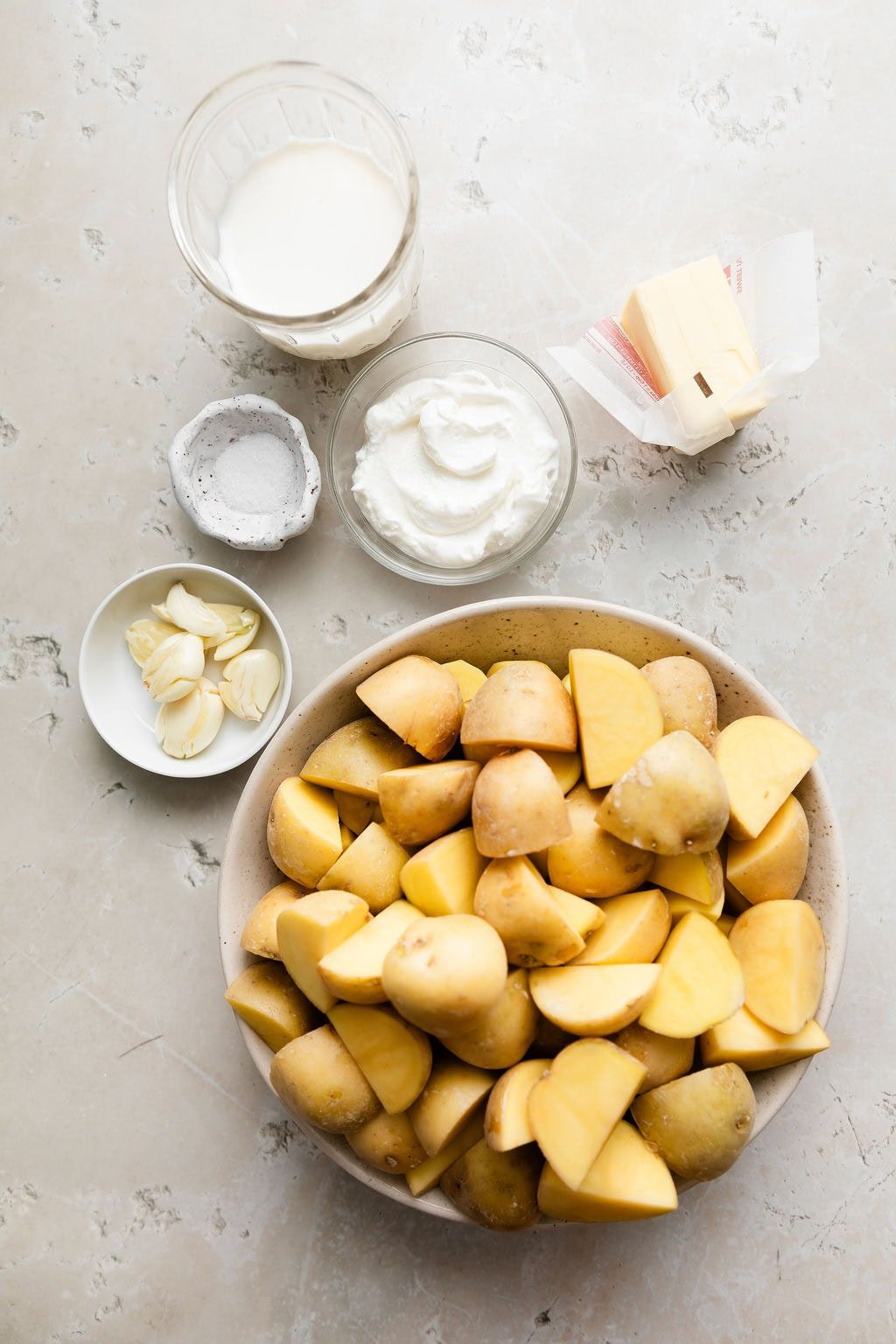 Overhead view of a variety of ingredients for Mashed Potatoes in different sized bowls on a marble countertop. 