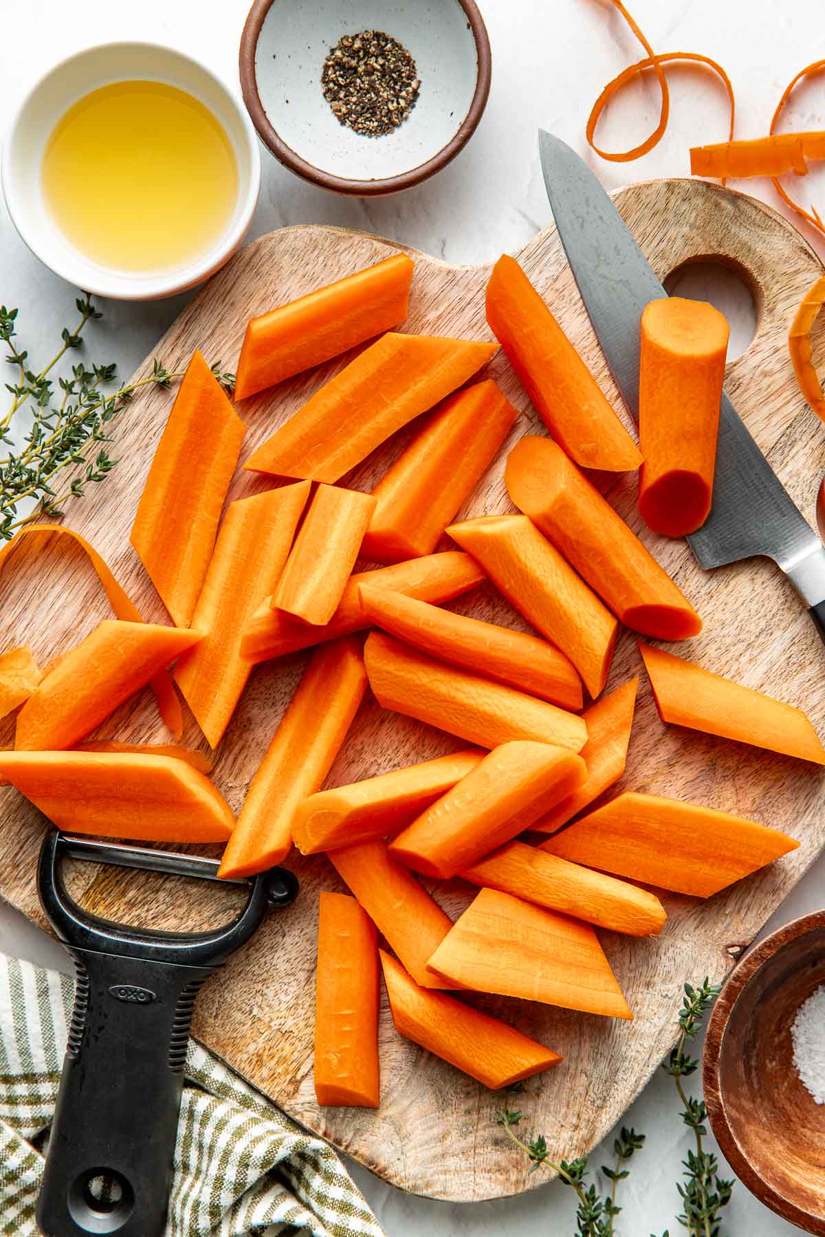 Overhead view of a wooden cutting board filled with peeling and cut carrots with olive oil and black pepper on the side in small bowls.