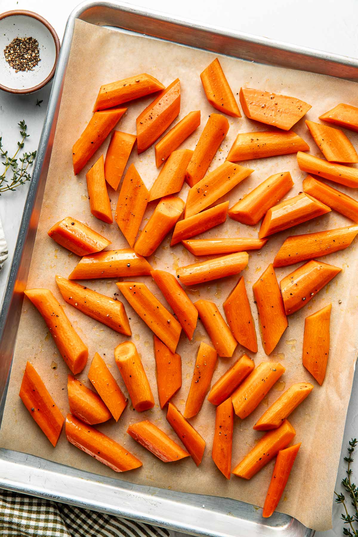 Overhead view of a sheet pan lined with parchment paper filled with peeled and cut carrots sprinkled with seasonings.