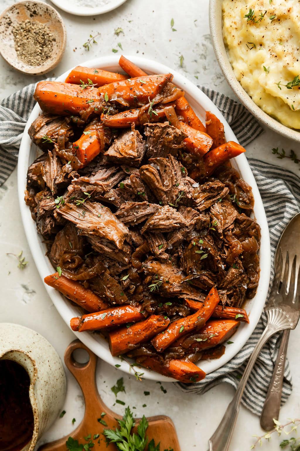 Close up view of an oval platter filled with shredded venison roast surrounded by carrots and topped with a brown gravy and sprinkled with fresh herbs and black pepper.