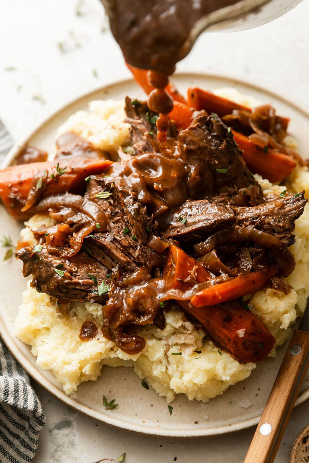 Close up view of a plate filled with venison roast and mashed potatoes with a gravy boat overhead pouring gravy on the mixture. 