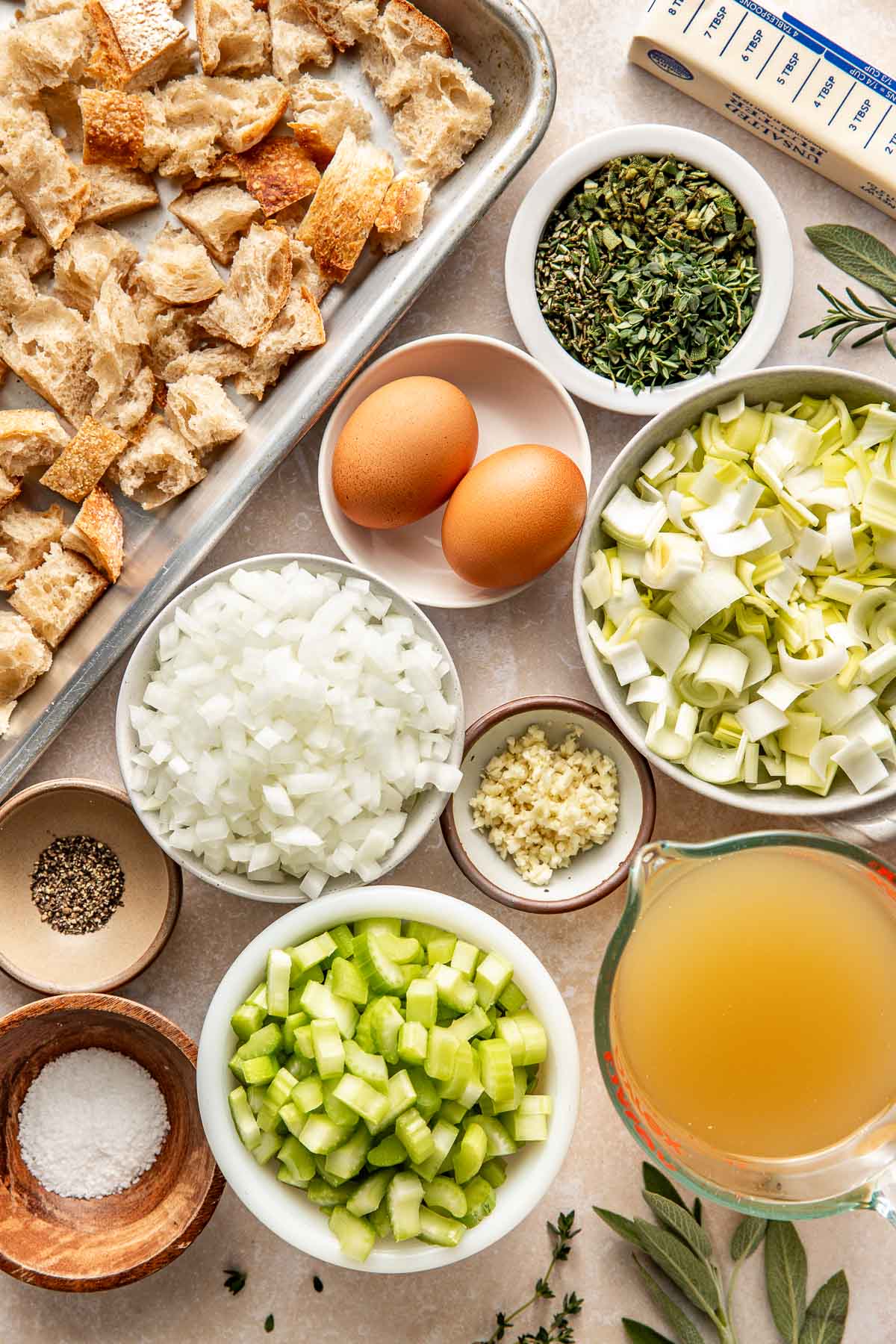 Overhead view of a variety of ingredients for stuffing in different sized bowls and a sheet pan filled with pieces of bread on the side.