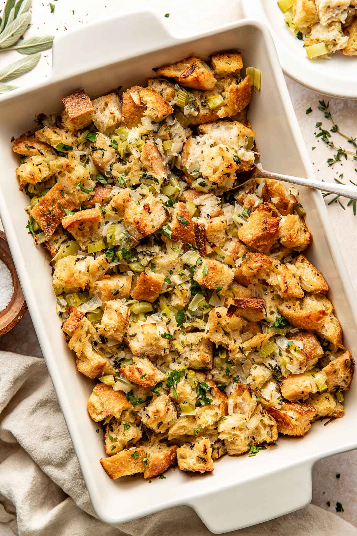 Overhead view of a white baking dish filled with stuffing topped with fresh herbs and black pepper.