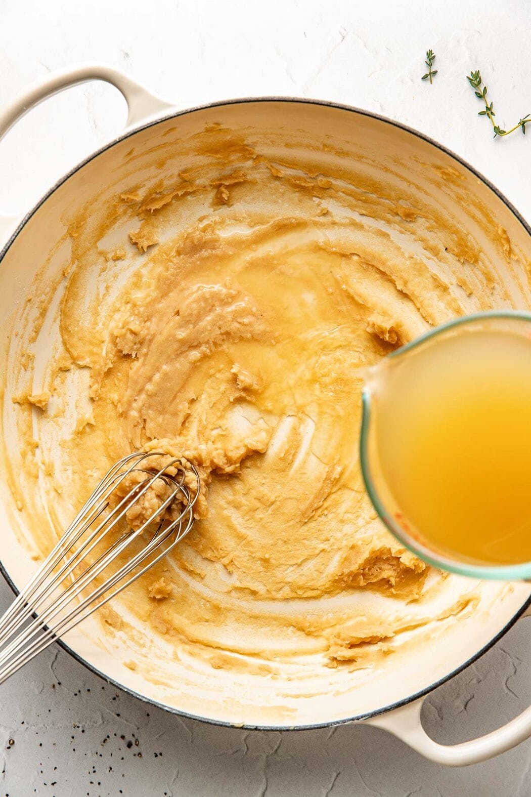 Overhead view of a white skillet with a roux being whisked with a wire whisk and broth being poured over top.