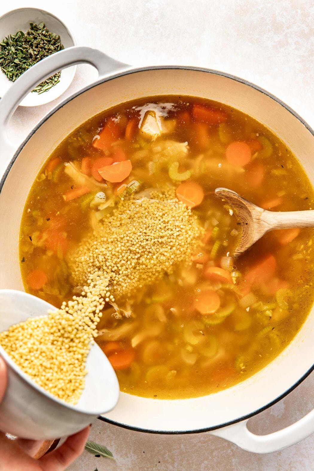 Overhead view of a Dutch oven filled with Turkey Soup and a hand pouring acini di pepe into the mixture. 