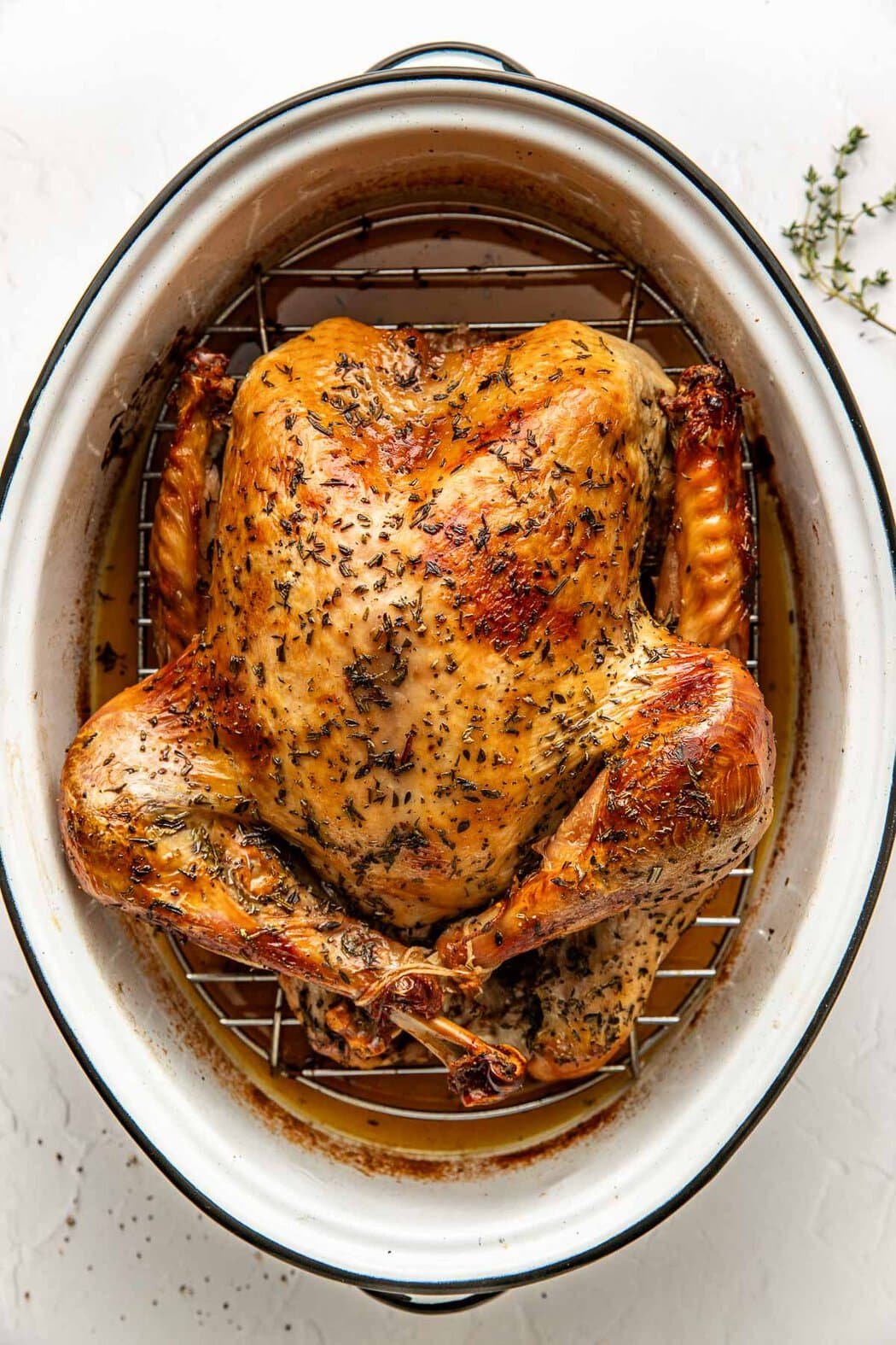 Overhead view of a freshly roasted turkey in a white roasting pan on a wire rack surrounded by drippings. 