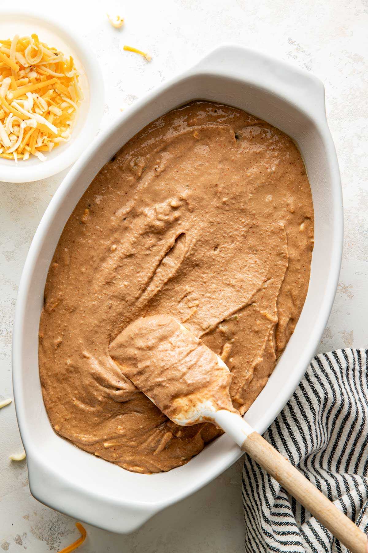 Overhead view of a white oval baking dish filled with Homemade Bean Dip mixture being spread with a spatula for a smooth top.