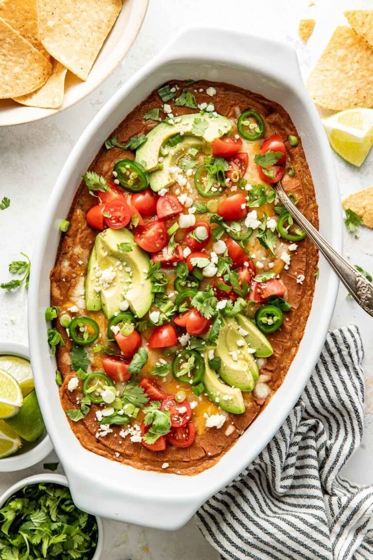 Overhead view of a white oval baking dish filled with homemade bean dip topped with fresh cilantro, diced tomatoes, and sliced jalapenos.