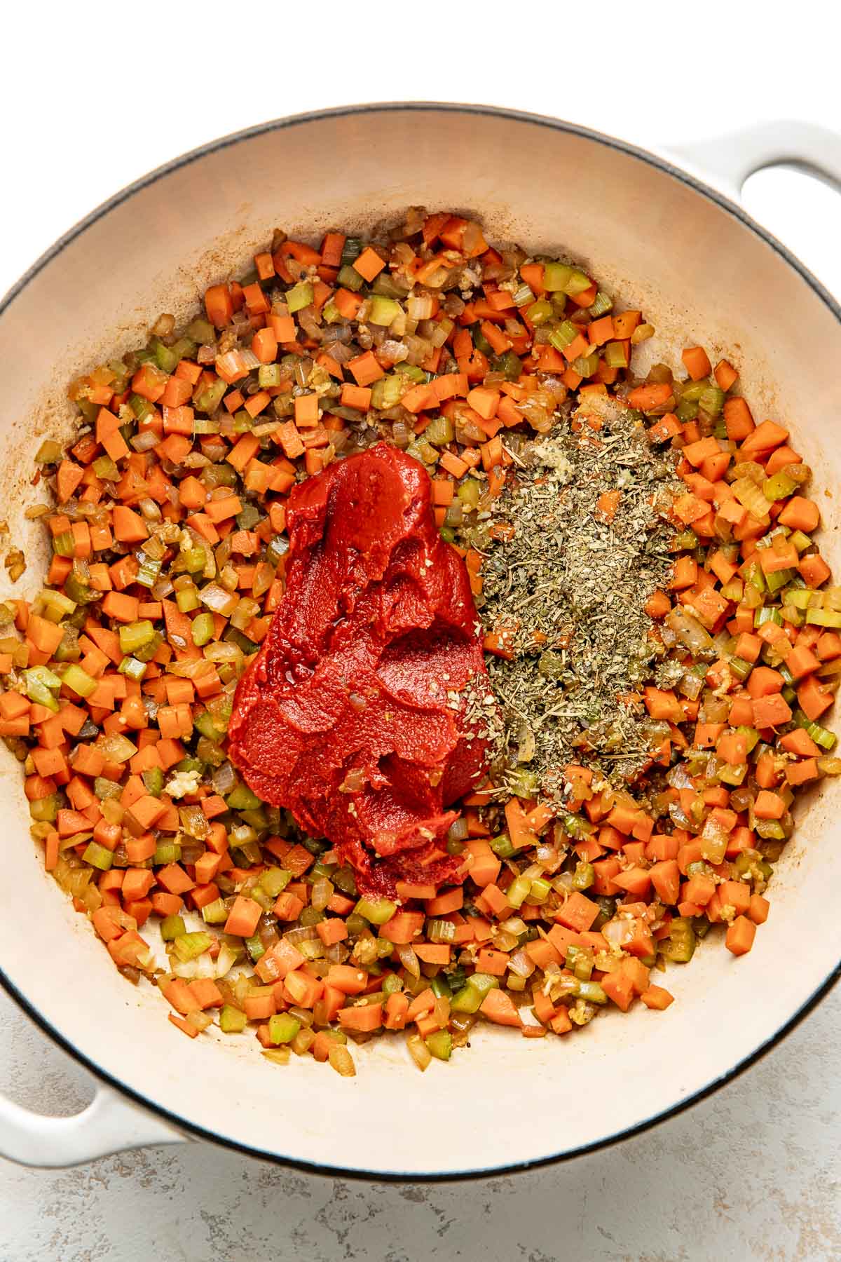 Overhead view of a white skillet filled with chopped vegetables, tomato paste, and dried herbs.