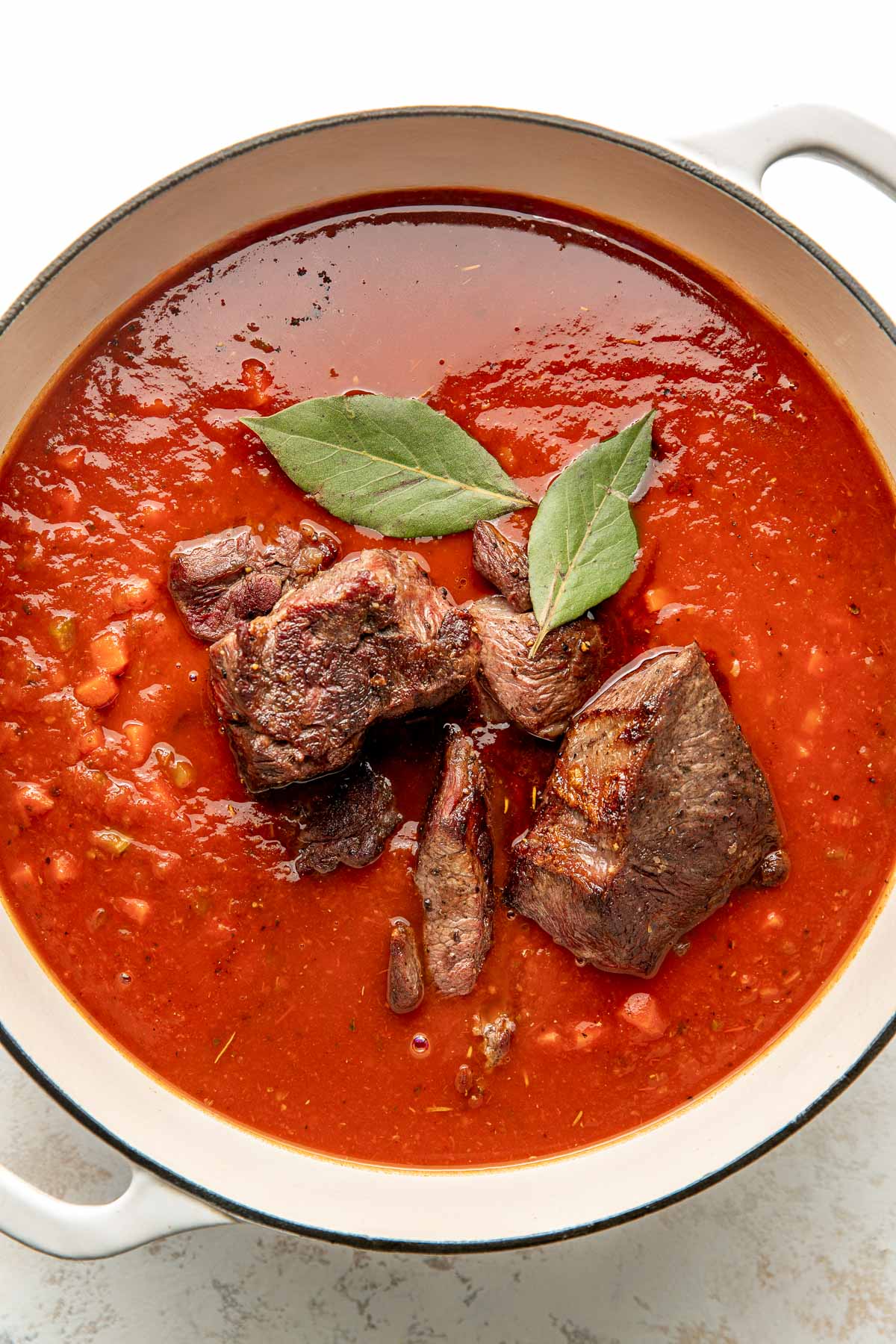 Overhead view of a white skillet filled with tomato sauce, chunks of seared beef, and two bay leaves.