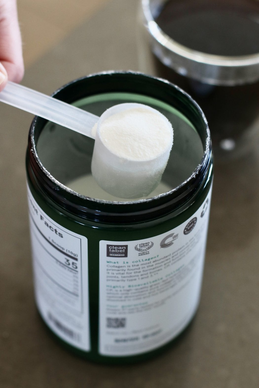 Overhead view of a canister of collagen with a hand bringing a scoop out showing the white powdery texture.