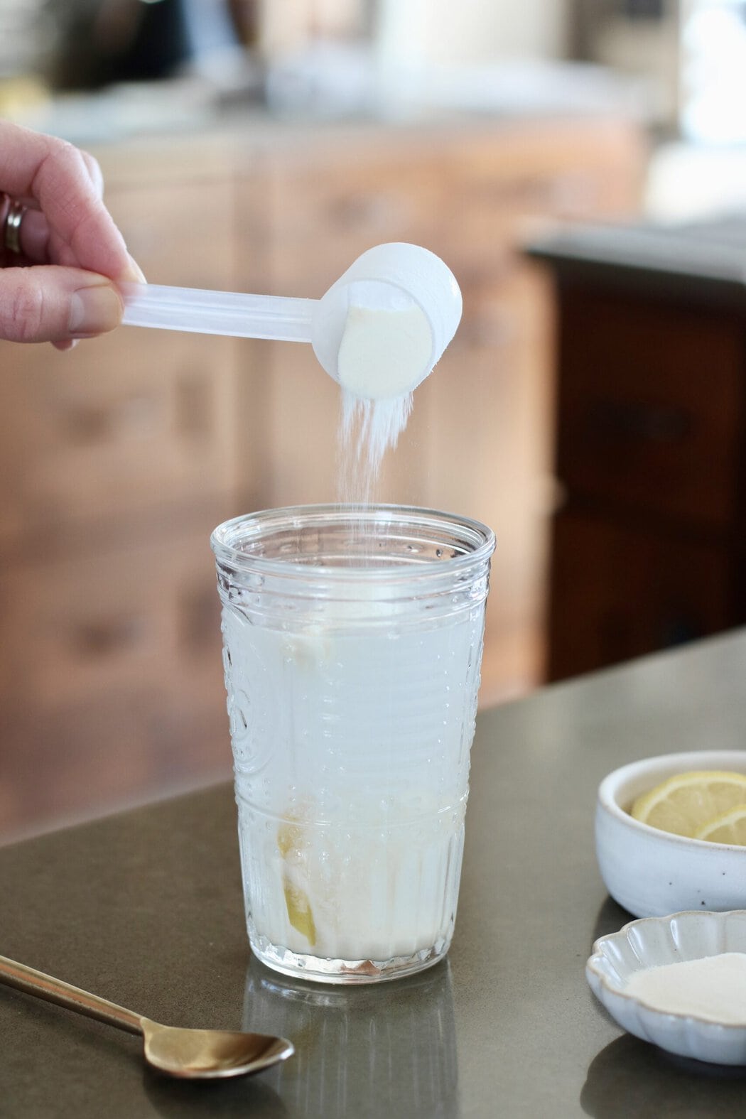 Close up view of a glass of water with a hand pouring a scoop of collagen into it.