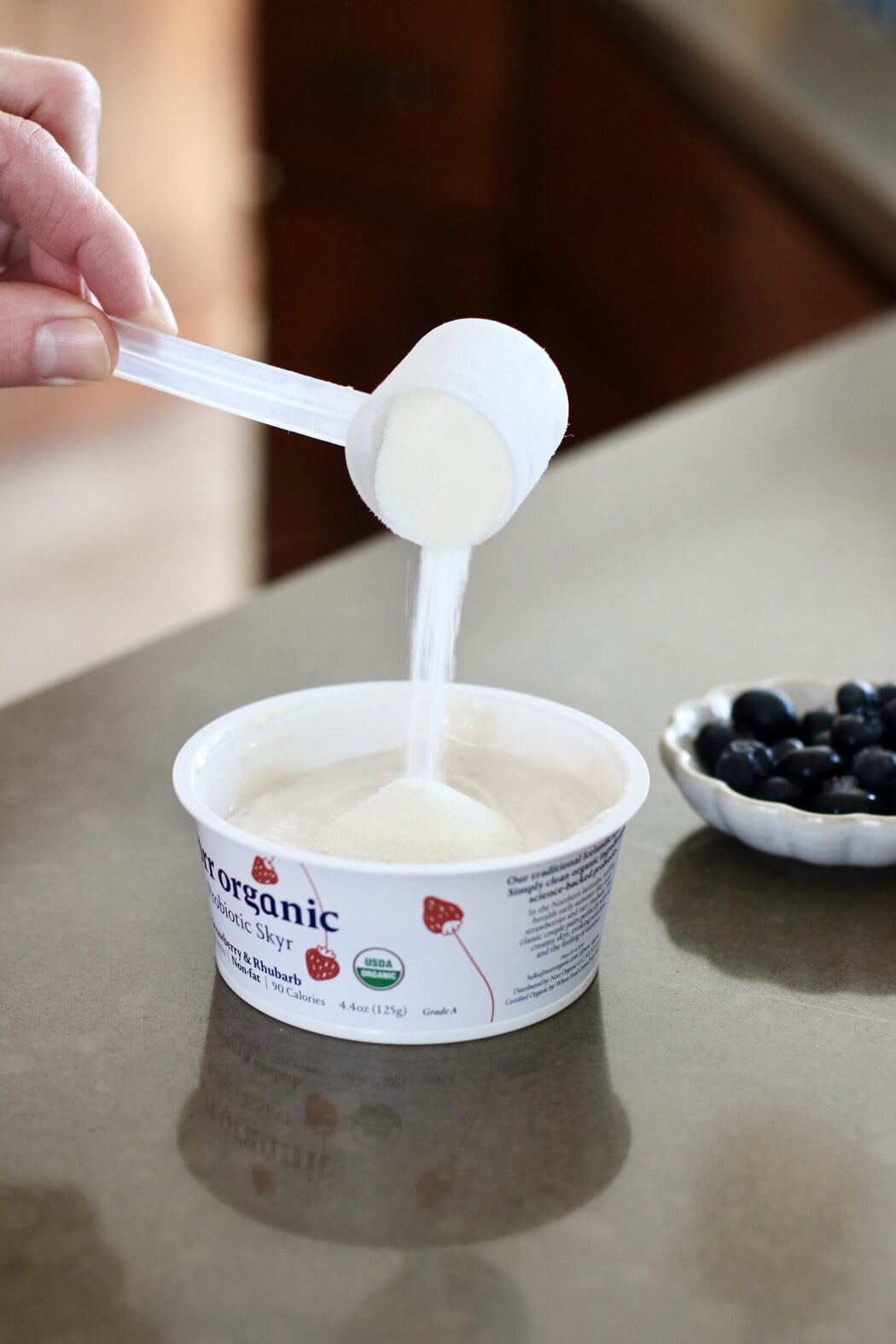 Close up view of a cup of yogurt on a kitchen countertop with a spoon of collagen being poured on top.