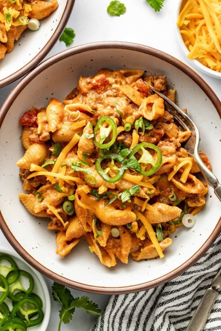 Overhead view of a serving of taco pasta in a white bowl garnished with shredded cheddar cheese, sliced jalapenos, and sliced green onions with a fork in the bowl.
