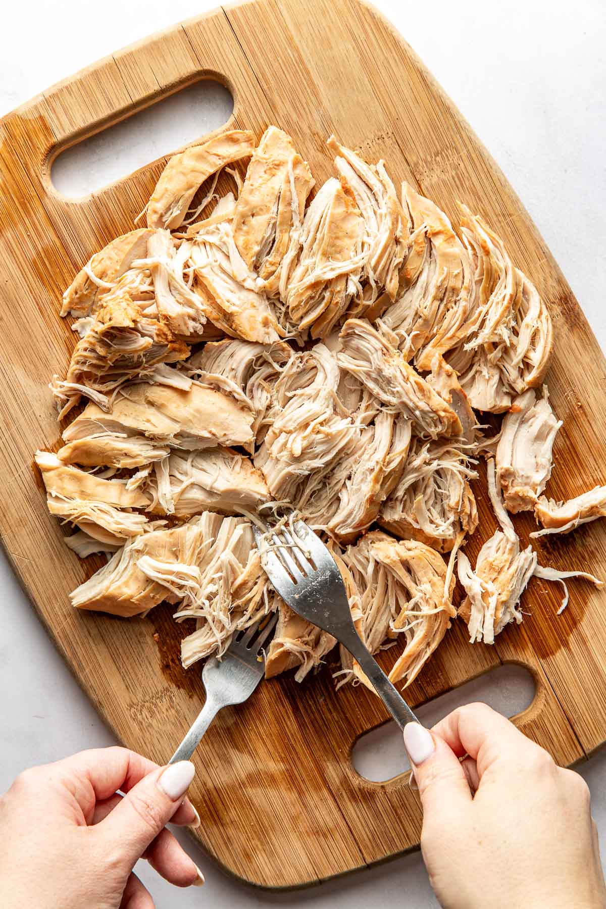Overhead view of a wooden cutting board filled with shredded chicken and two hands using forks to pull the meat apart.
