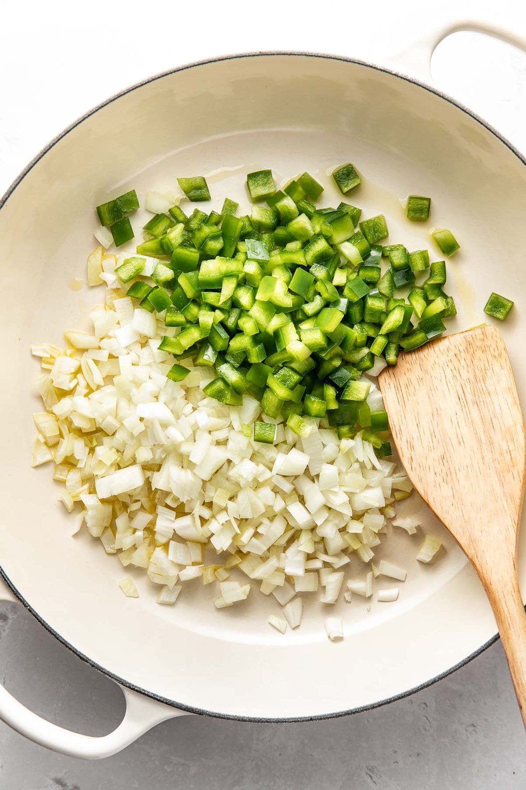 Overhead view of a white skillet filled with diced onions and green peppers and a wooden spatula on the side.