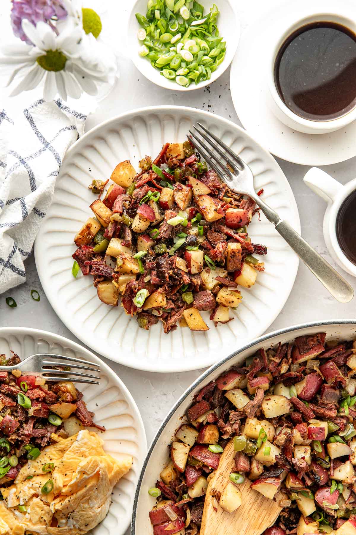 Overhead view of a tablescape with plates filled with Corned Beef Hash, white linen towels, and fresh cut green onions in a dish.