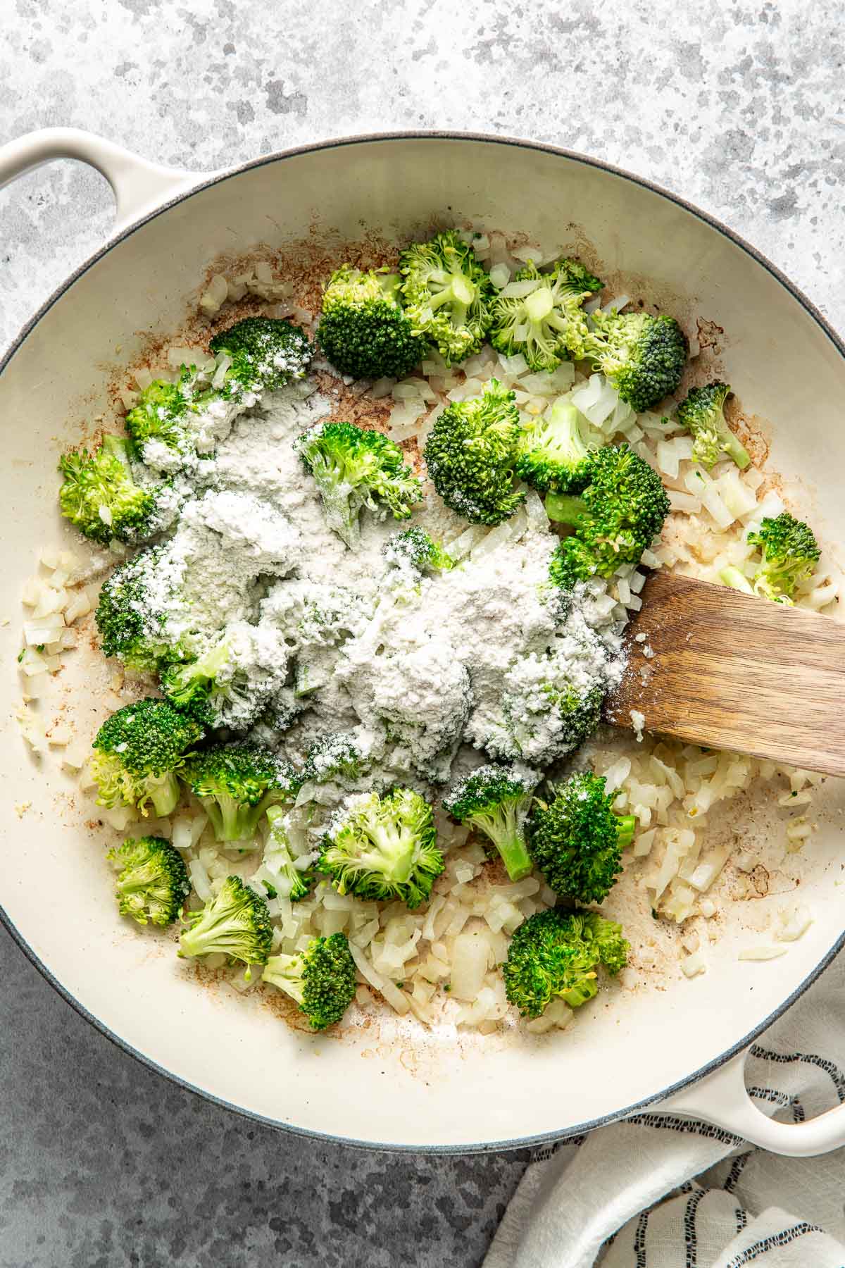 Overhead view of a skillet filled with onions and broccoli with flour over the top.