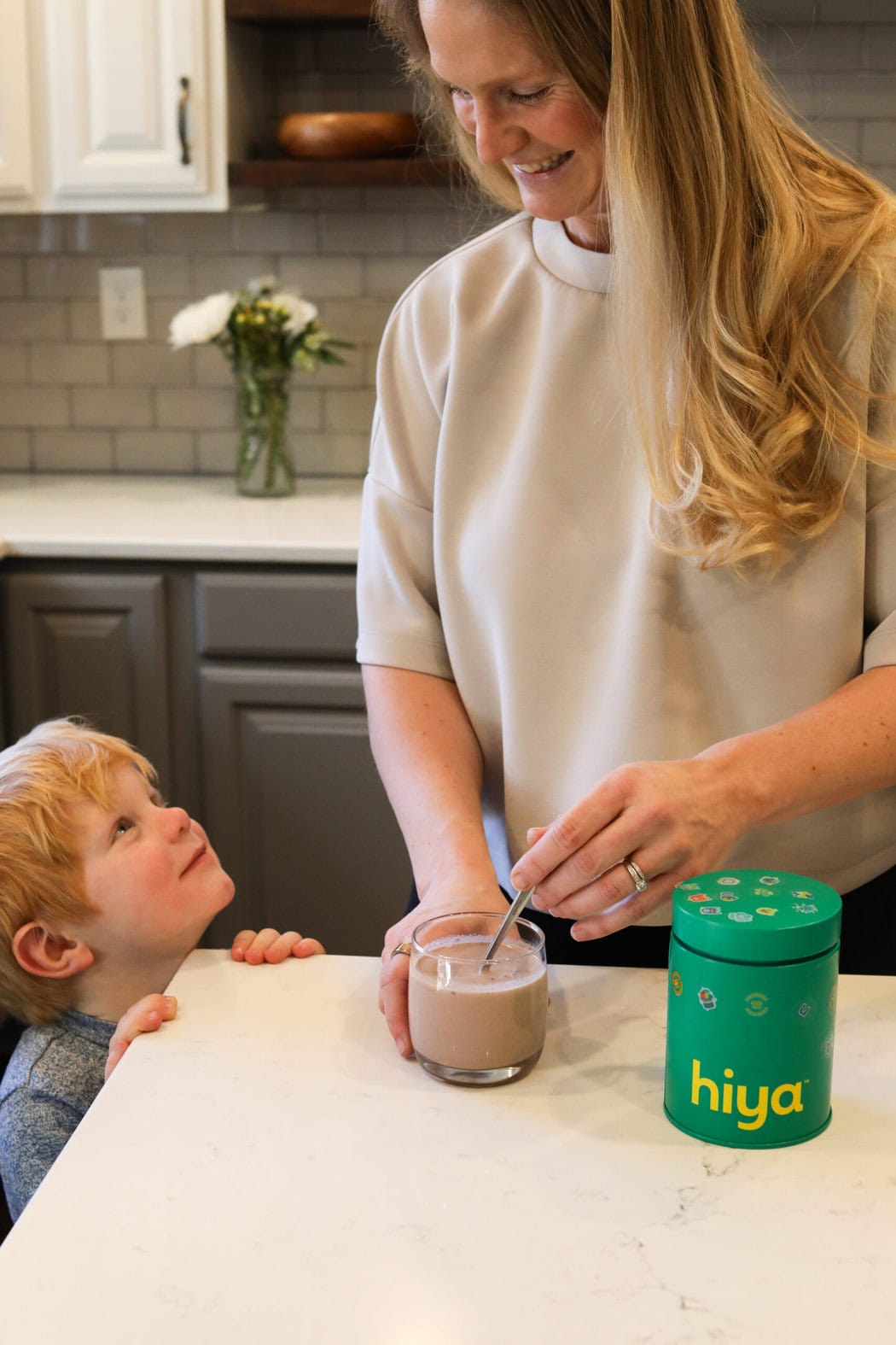 A woman and child at a kitchen counter mixing a glass of hiya drink mix with a spoon.