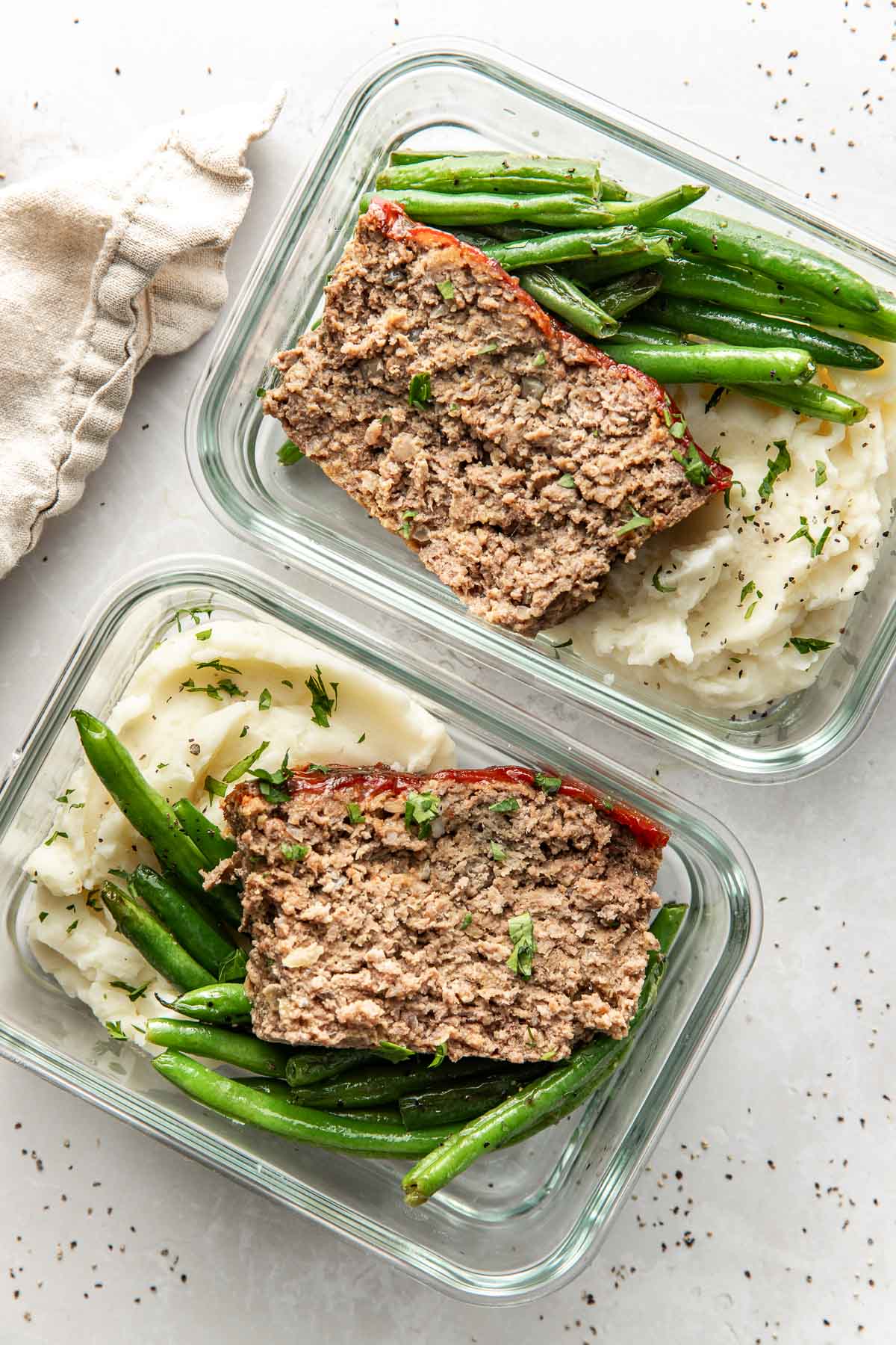 Overhead view of two glass meal prep containers filled with mashed potatoes, green beans, and meatloaf all topped with fresh herbs.