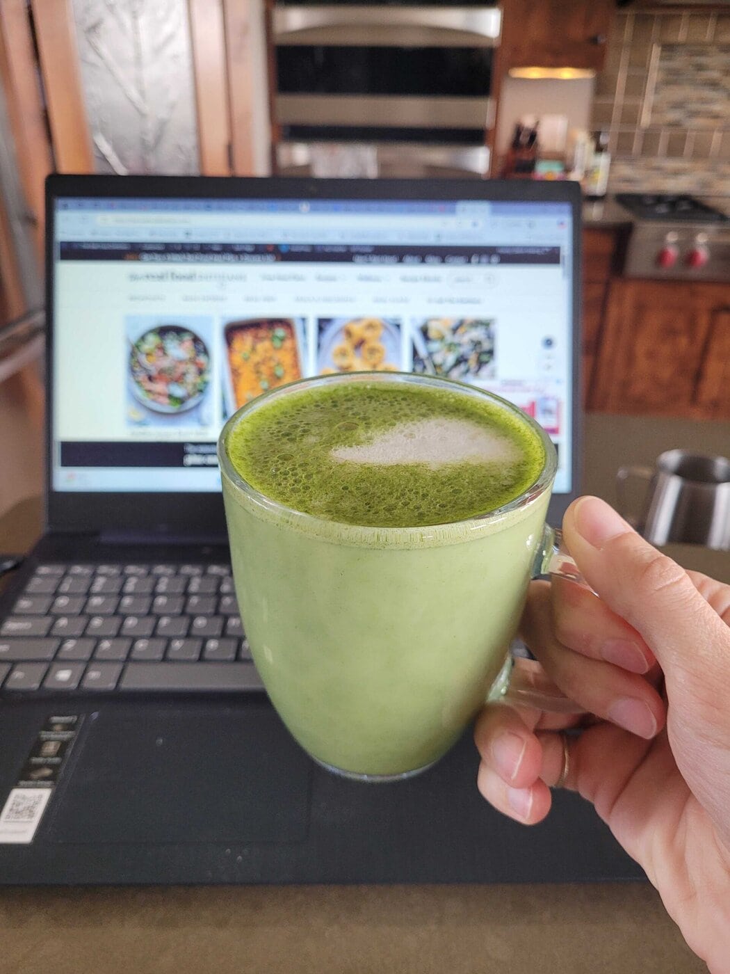 A hand holding a glass mug of a matcha latte with a frothy top.