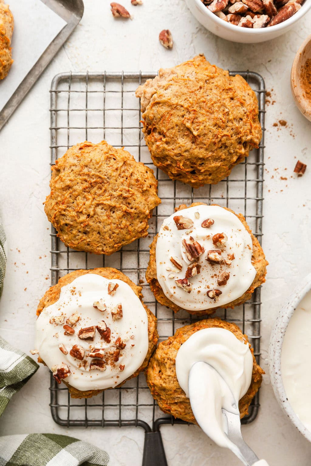Overhead view of a cooling rack of Carrot Cake Cookies being frosted with a cream cheese frosting and sprinkled with pecan pieces. 