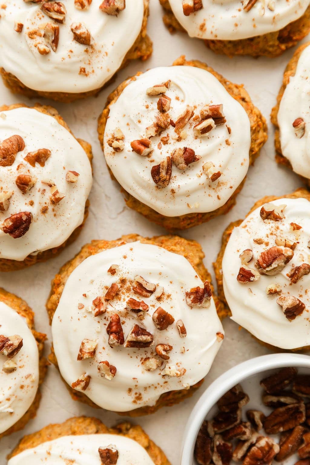 Overhead view of carrot cake cookies topped with cream cheese frosting and sprinkled with pecan pieces and cinnamon. 