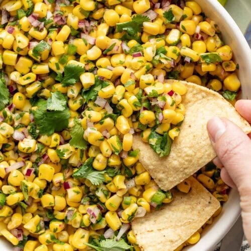 Overhead view of a bowl of Chipotle Corn Salsa with a hand scooping a tortilla chip into it.